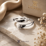 A polished silver ring with a sculptural, intertwined organic form, displayed on a textured beige stone slab next to a beige silk ribbon and a branded ZIRAavi box.