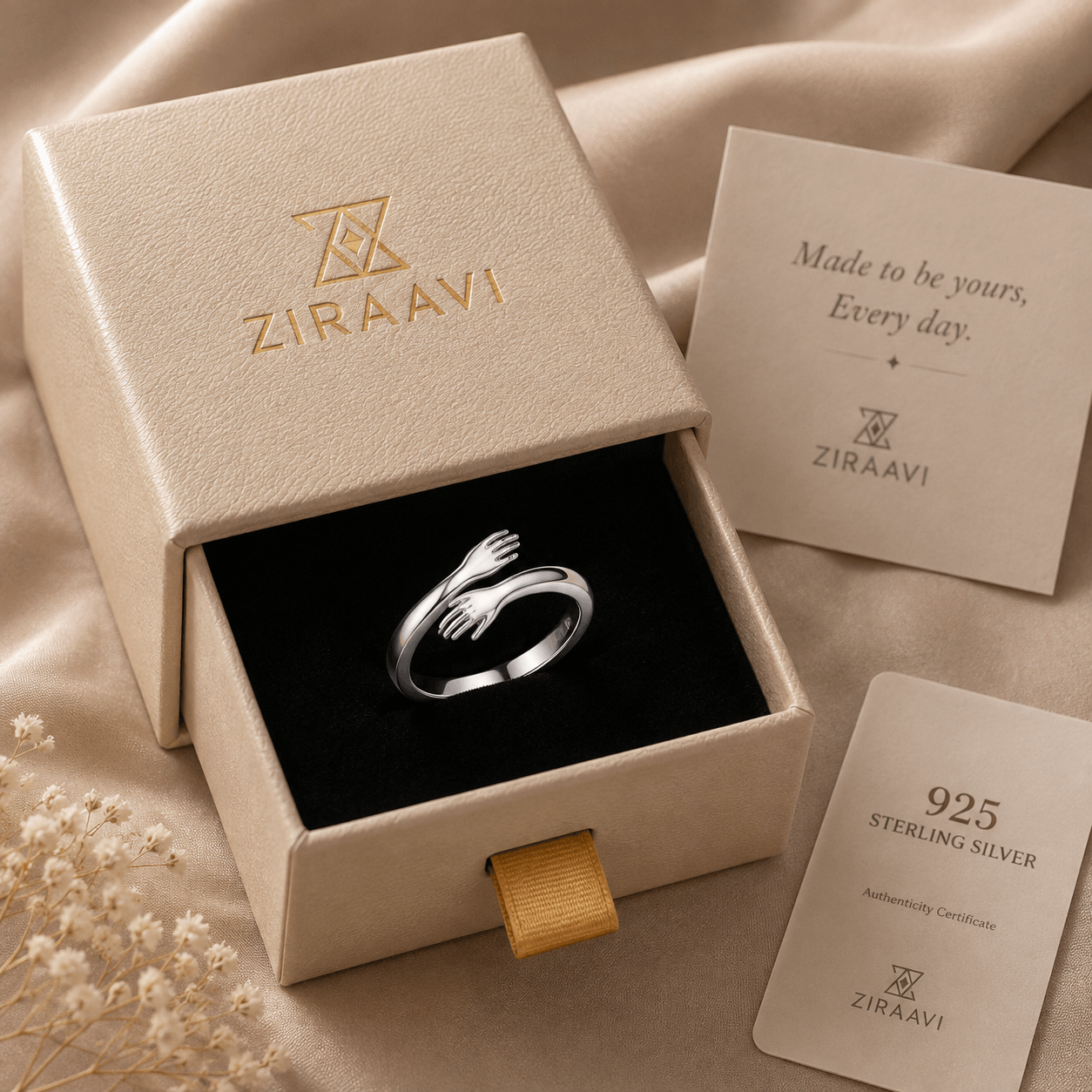 A silver ring with two interlocking hands is displayed inside an open beige ZIRAAVI jewelry box on a black velvet insert. The box lid is printed with the ZIRAAVI logo, and two branded cards are placed beside it on a beige satin fabric background.