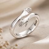 A polished sterling silver ring featuring two interlocking hand designs, resting on a soft beige fabric background with dried flowers in the corner. The inner band is stamped with 'S925'.
